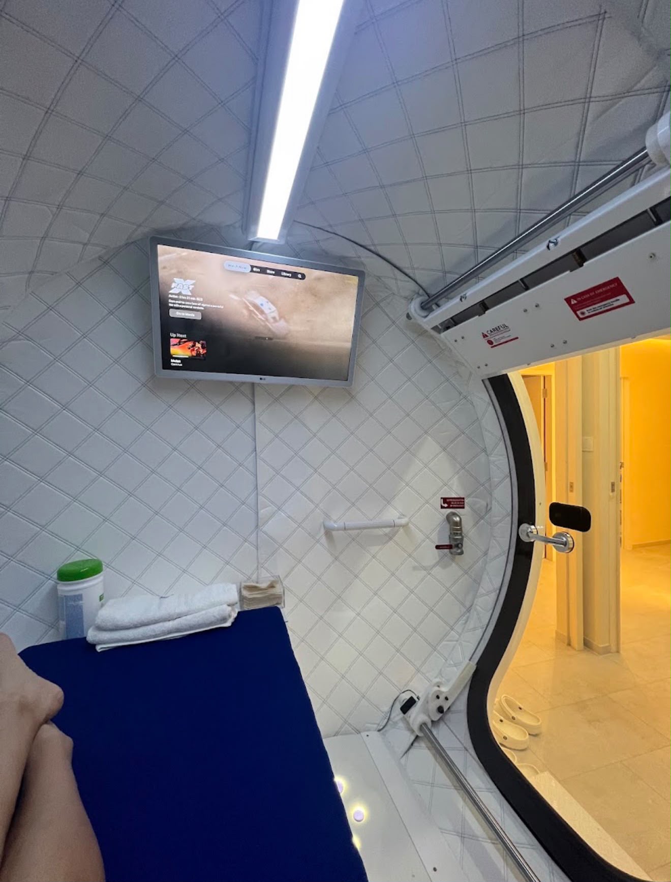 Inside the hyperbaric oxygen chamber at The Longevity Science Centrum, with a mounted entertainment screen above the reclining bed and towels laid out.
