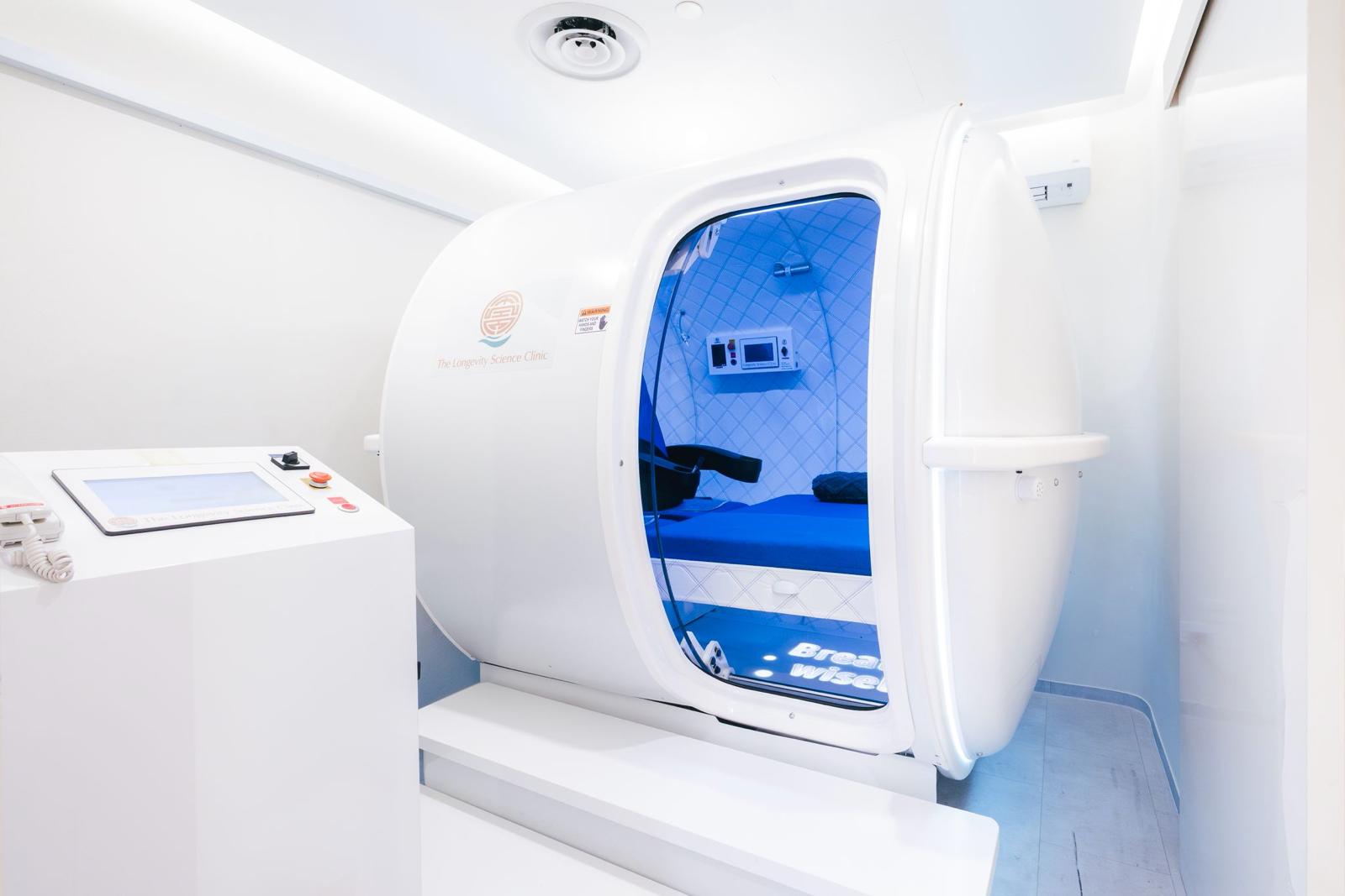The white hyperbaric oxygen chamber carrying The Longevity Science Centrum emblem, beside its control console.