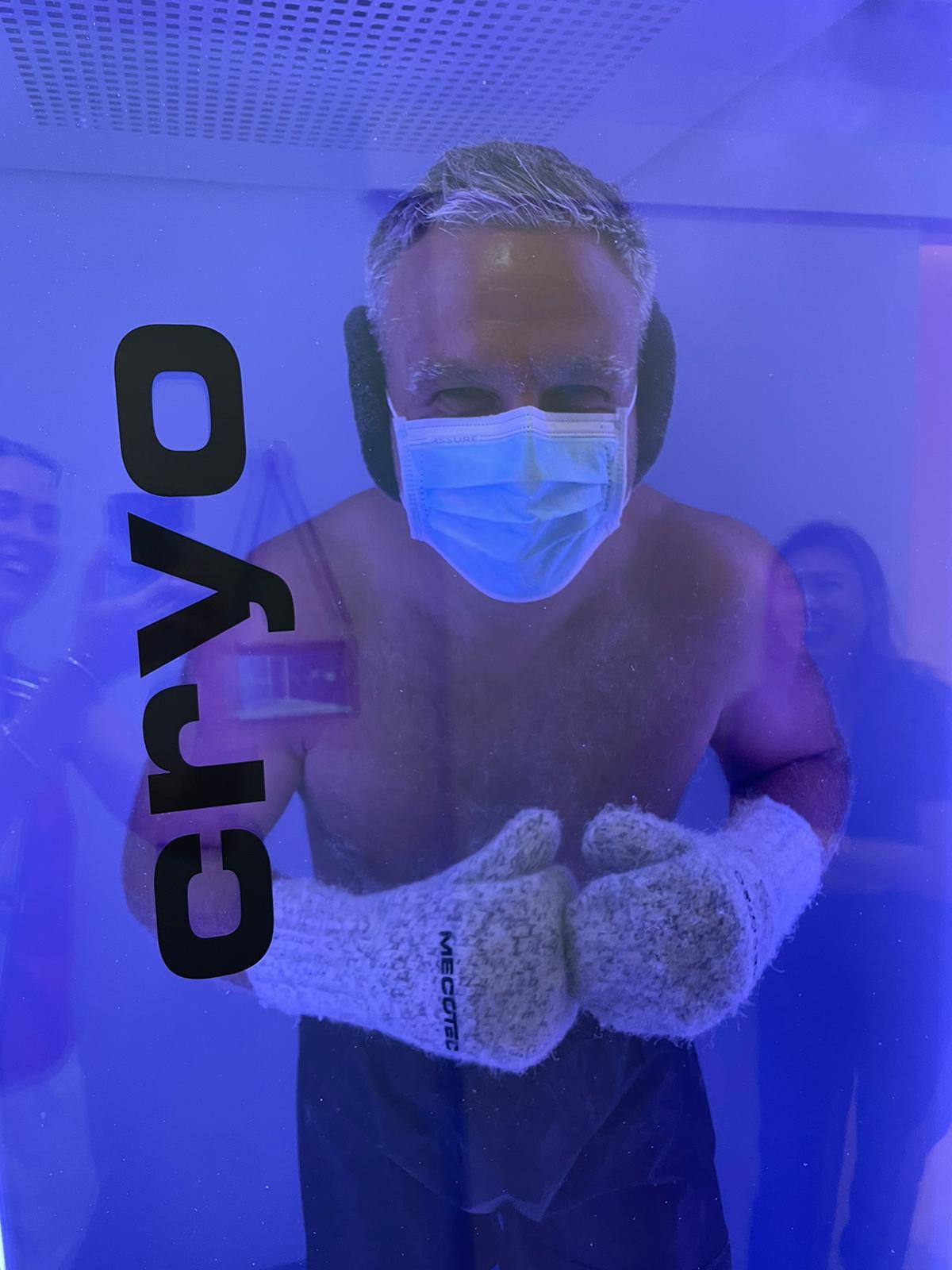A guest smiling inside the whole-body cryotherapy chamber at The Longevity Science Centrum, wearing a face mask, earmuffs and protective gloves in the blue-lit cabin.