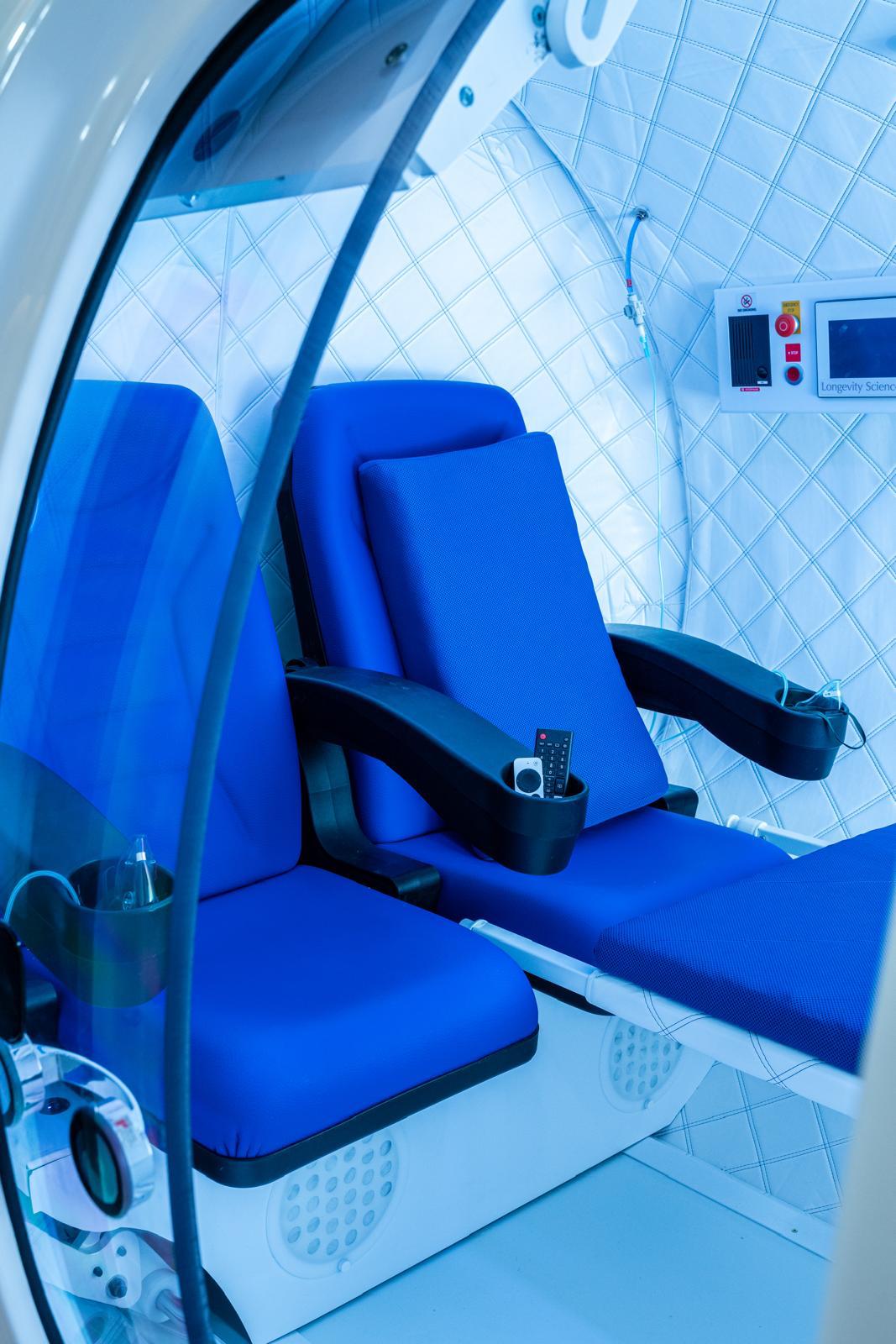 Inside the hyperbaric oxygen chamber at The Longevity Science Centrum, two blue seats and a reclining bed lit in cool blue light.