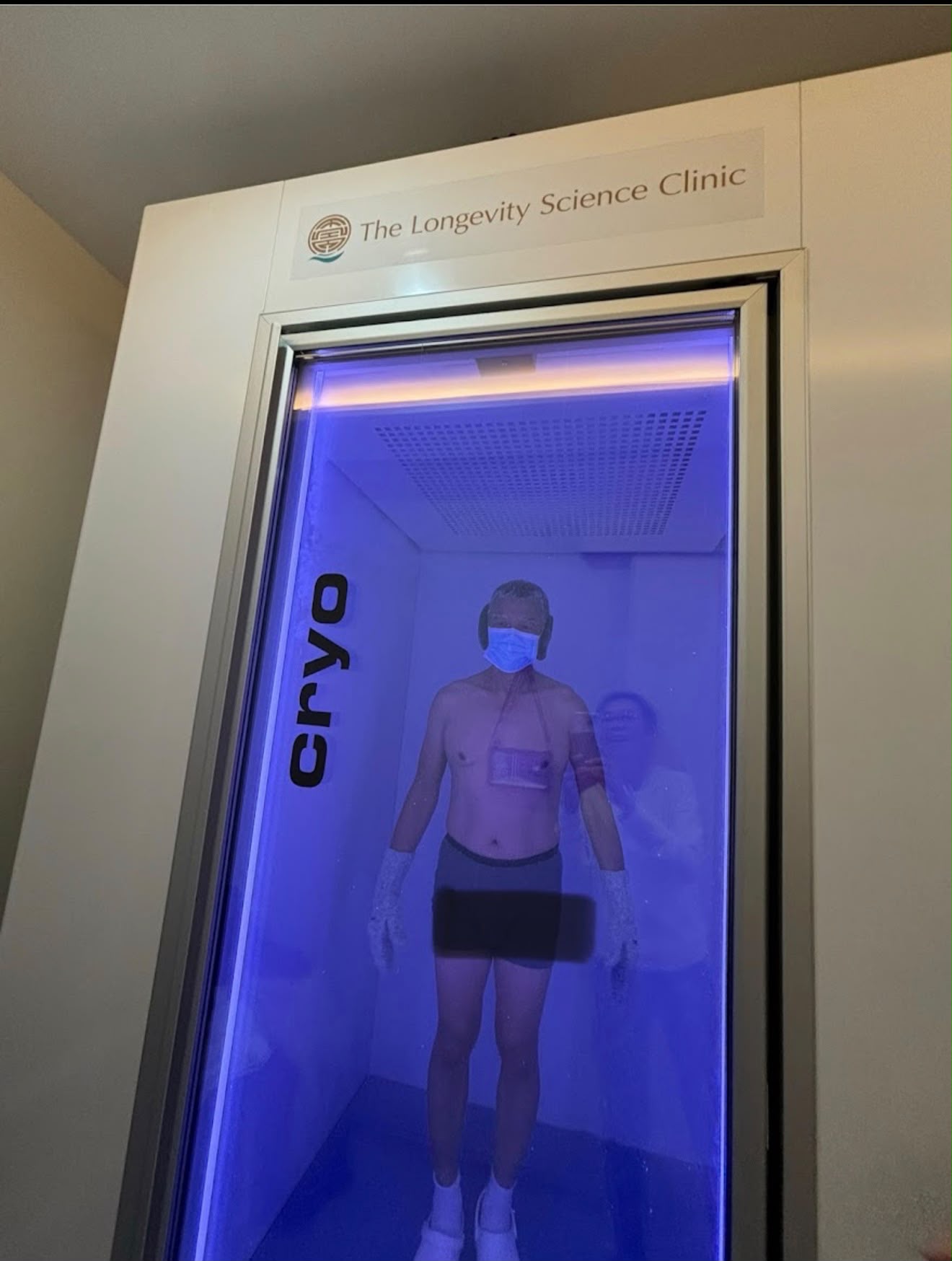 A guest standing inside the whole-body cryotherapy chamber at The Longevity Science Centrum, seen through the blue-lit glass door.