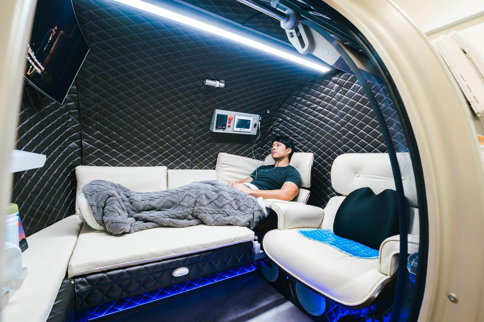 A client resting inside the hyperbaric oxygen chamber at The Longevity Science Centrum, wearing an oxygen cannula on a white leather recliner.